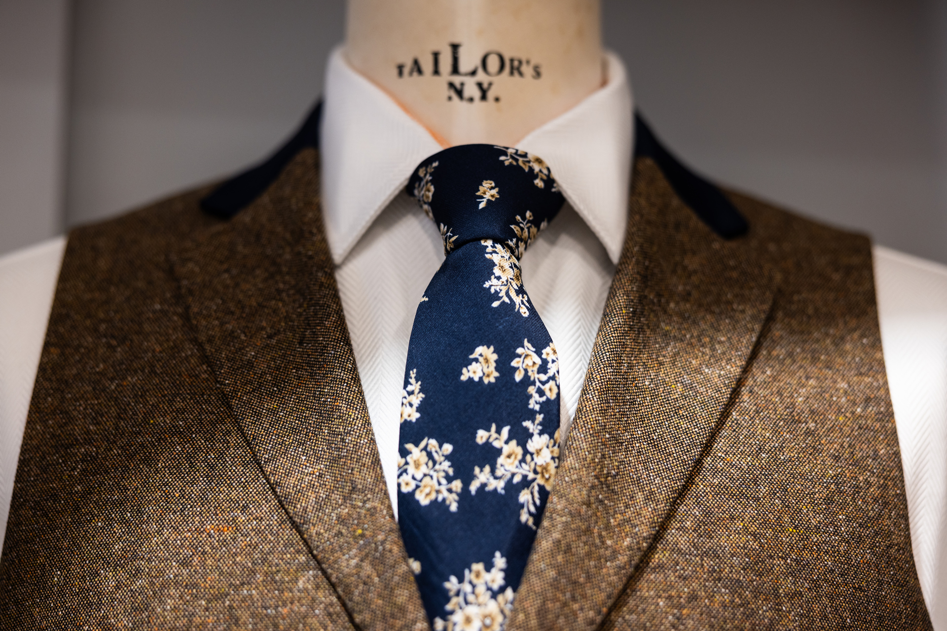 Close-up of a tweed jacket with a navy floral tie and crisp white shirt on a tailor's mannequin