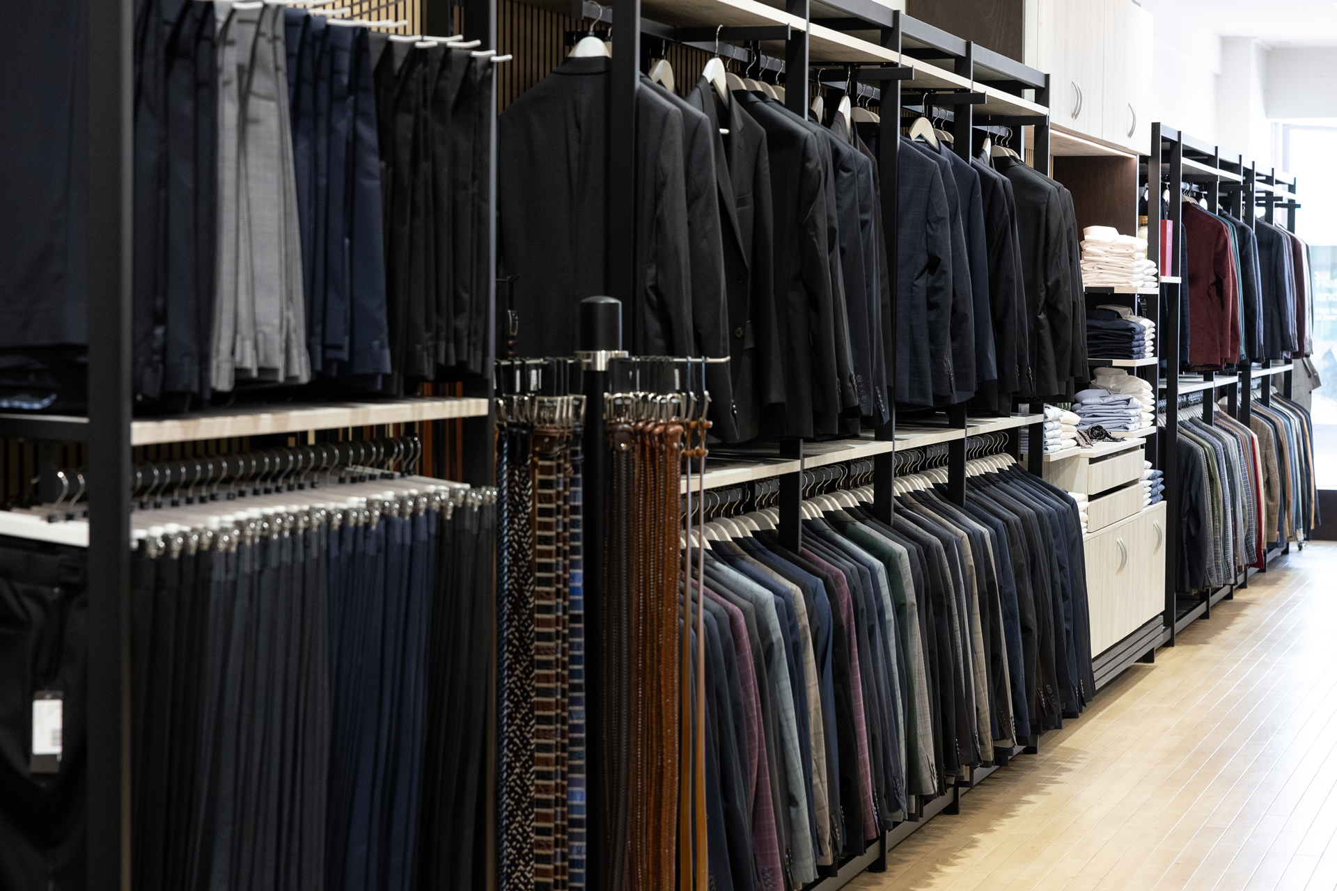 A long row of suit jackets, trousers, and belts hanging on racks along the store's shelving wall